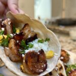 A close-up of a hand holding a taco filled with grilled shrimp, corn, fresh cilantro, and crumbled cheese in a lightly toasted tortilla.