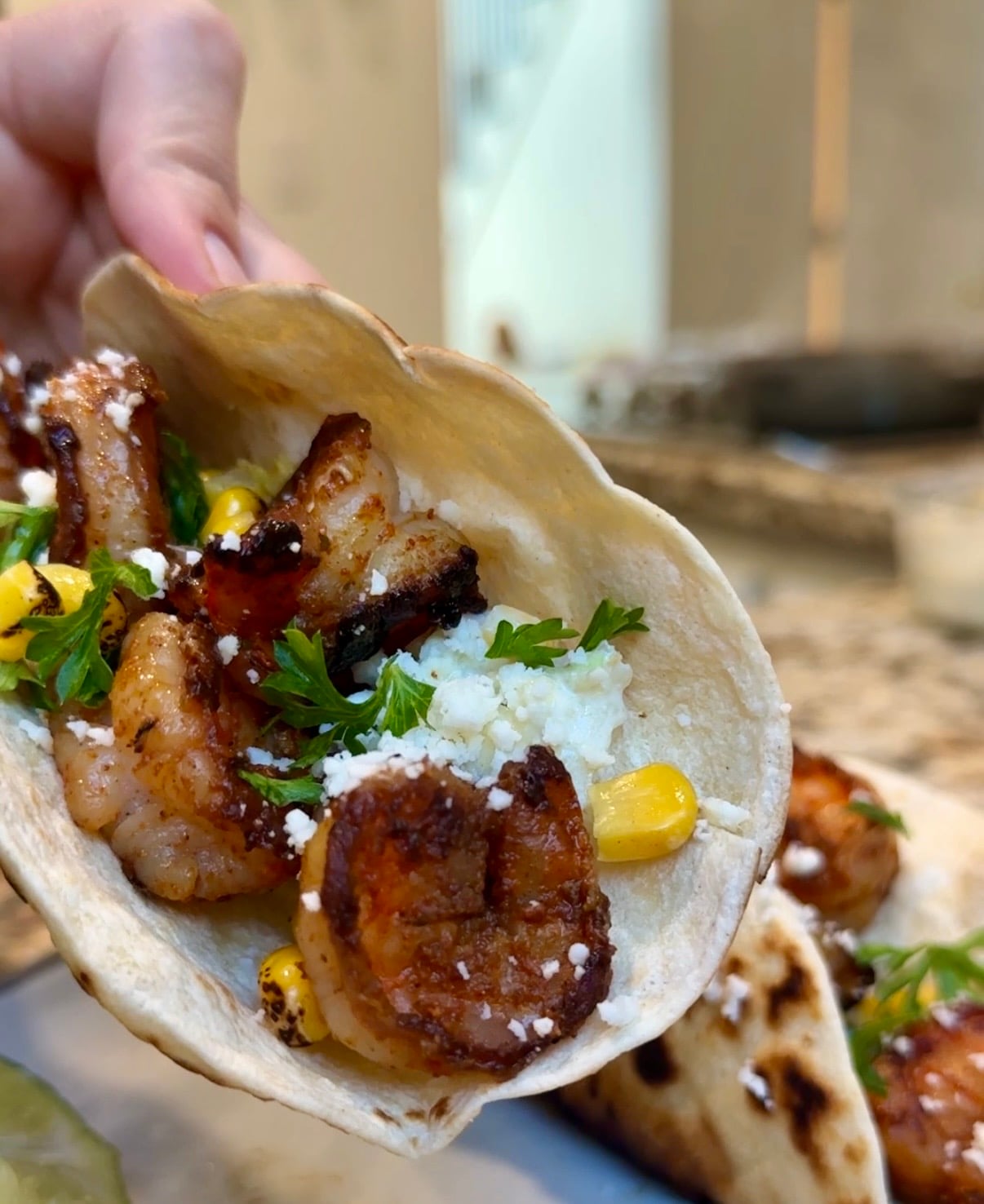 A close-up of a hand holding a taco filled with grilled shrimp, corn, fresh cilantro, and crumbled cheese in a lightly toasted tortilla.