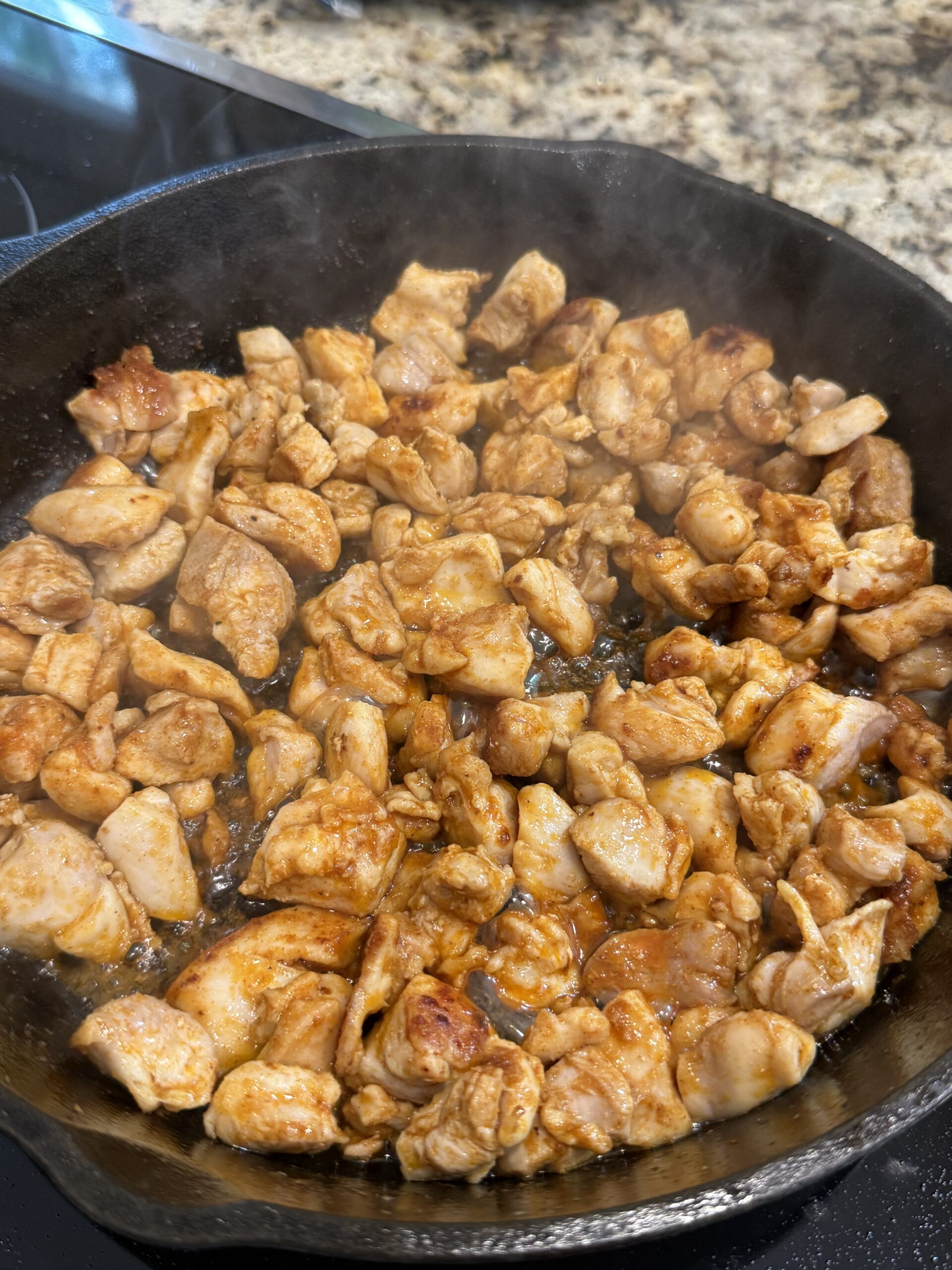 Diced, seasoned chicken pieces are sizzling in a black cast iron skillet on a stovetop, with steam rising and a granite countertop visible in the background.