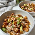 Two white bowls filled with a dish of roasted sweet potatoes, black beans, melted cheese, barbecue sauce, diced avocado, and chopped green onions on a white table. A napkin and plates are in the background.