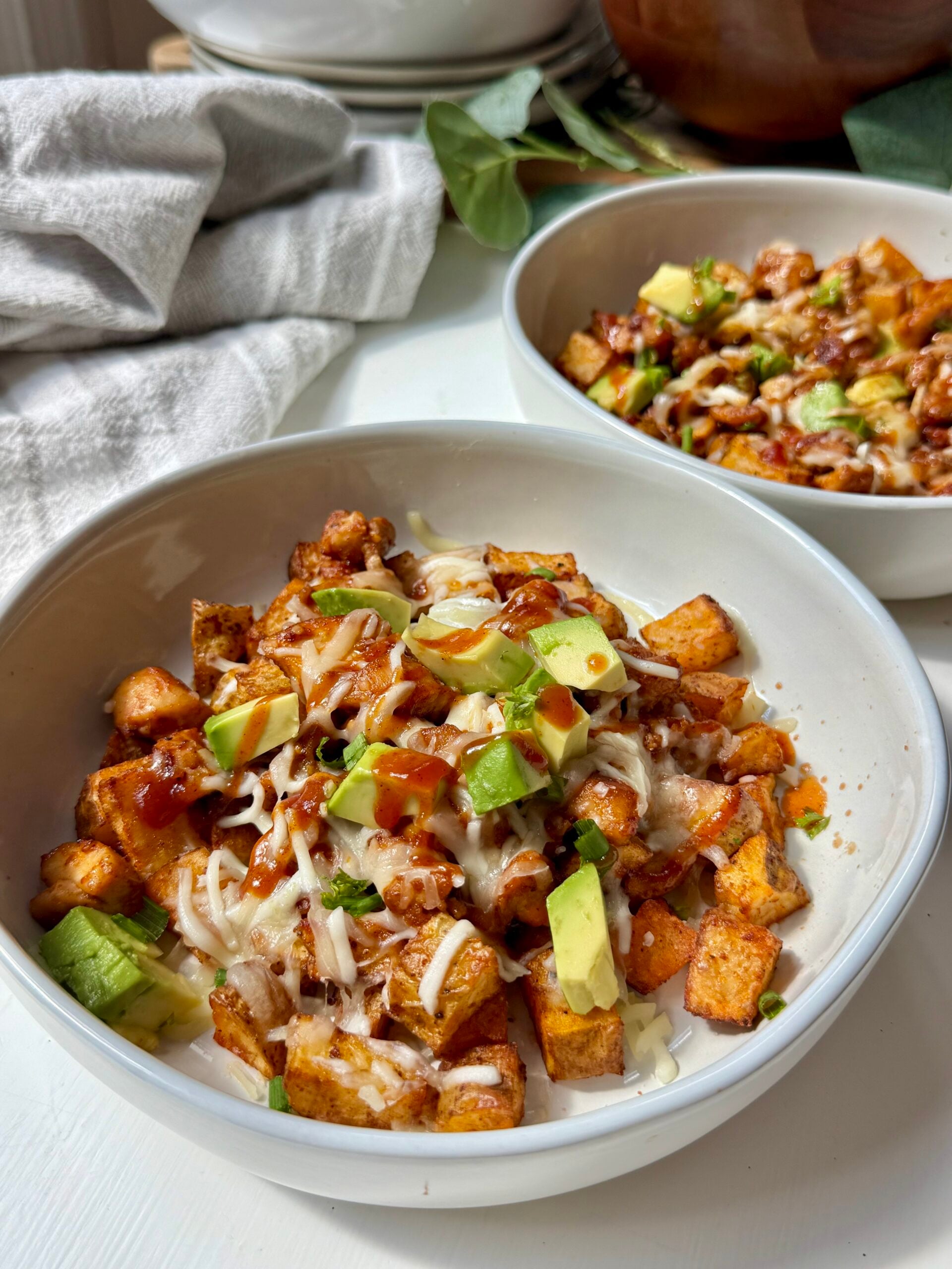 Two white bowls filled with a dish of roasted sweet potatoes, black beans, melted cheese, barbecue sauce, diced avocado, and chopped green onions on a white table. A napkin and plates are in the background.