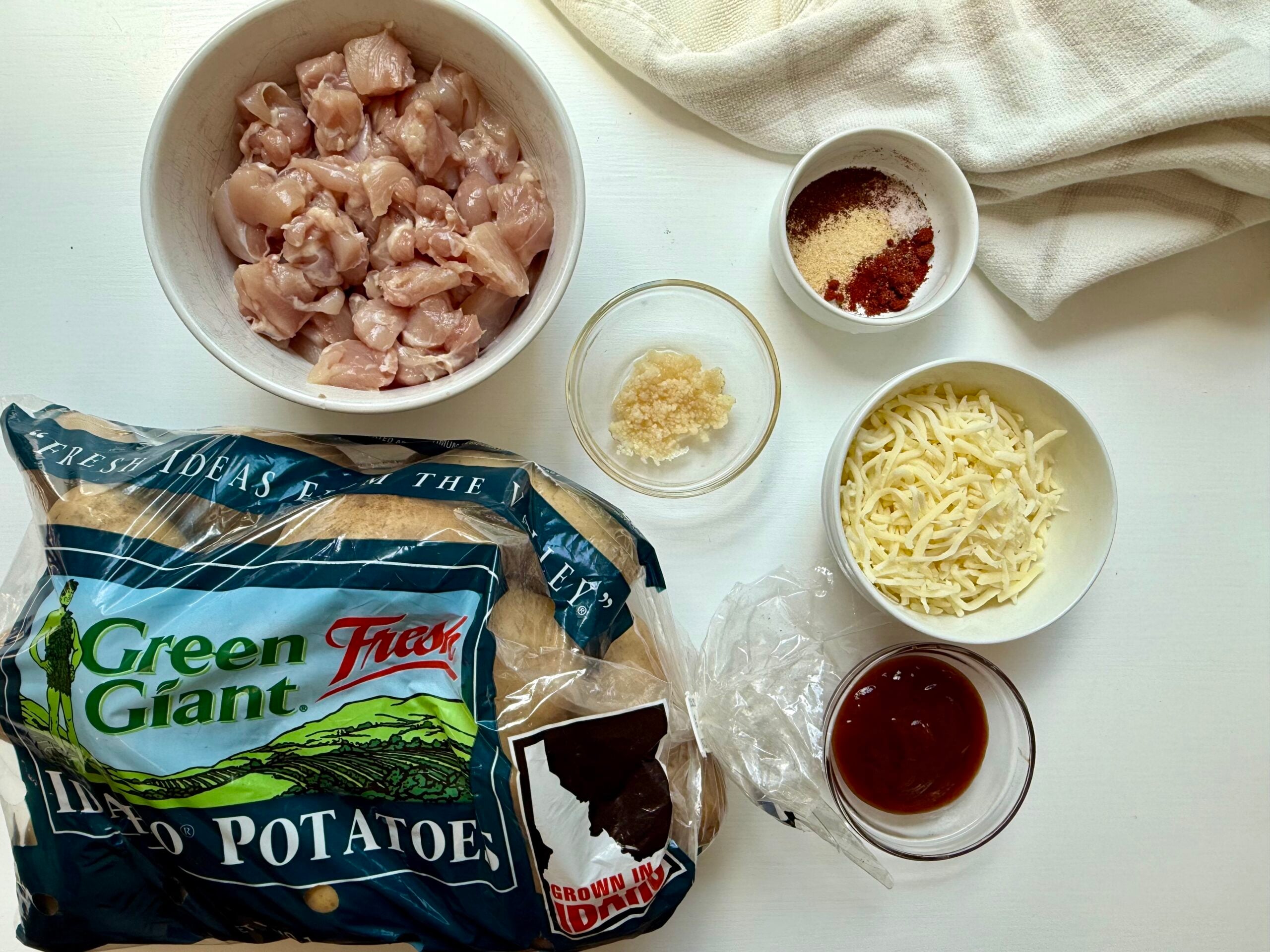 Ingredients on a white table: diced raw chicken, shredded cheese, minced garlic, barbecue sauce, seasonings, and a bag of Green Giant Fresh Idaho potatoes, with a white cloth in the corner.