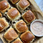 A tray of golden-brown slider sandwiches with sesame seed buns, melted cheese, and beef, served with a small bowl of creamy dipping sauce on parchment paper.