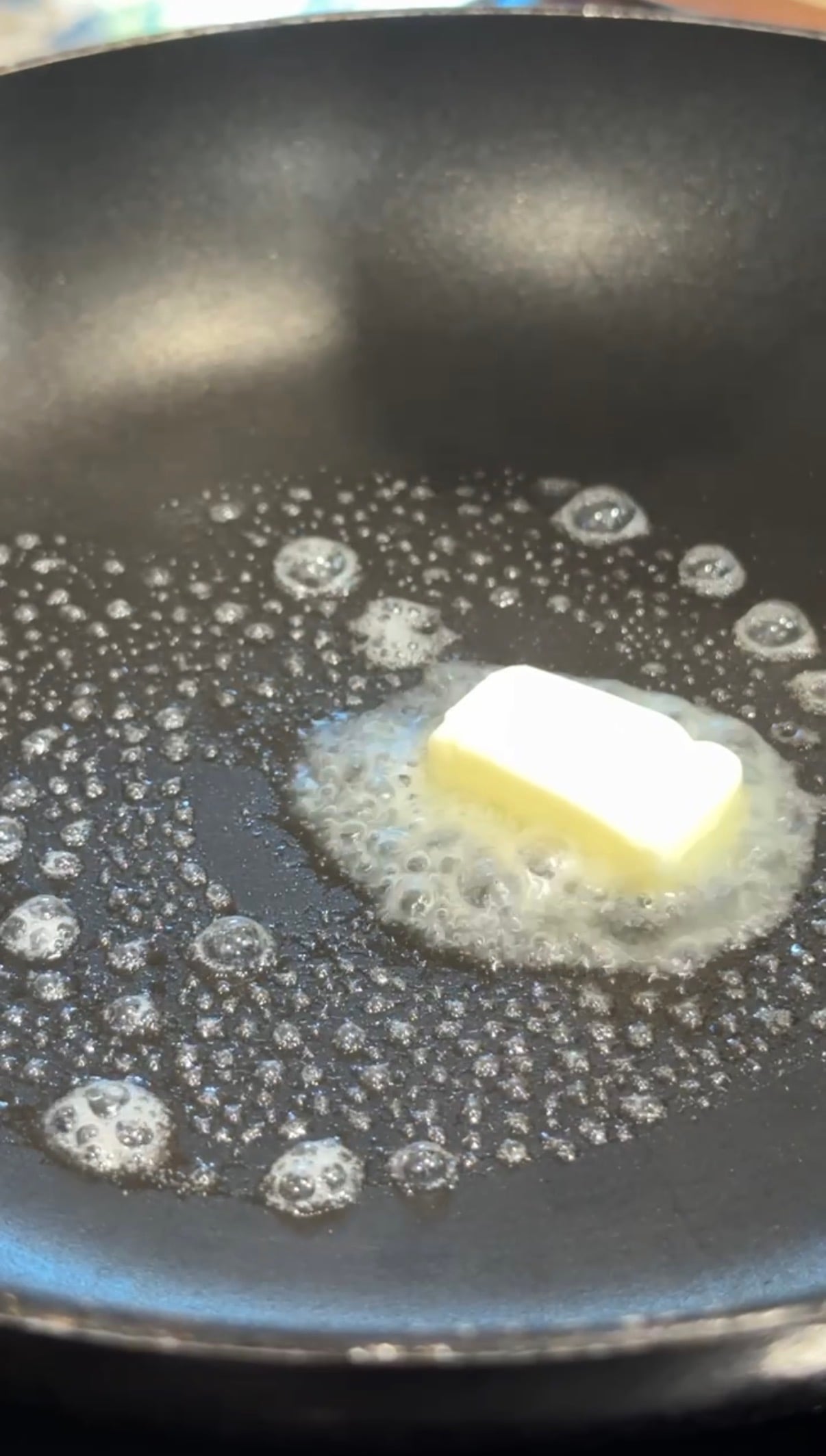 A pat of butter melting and bubbling in a black frying pan, surrounded by sizzling foam.