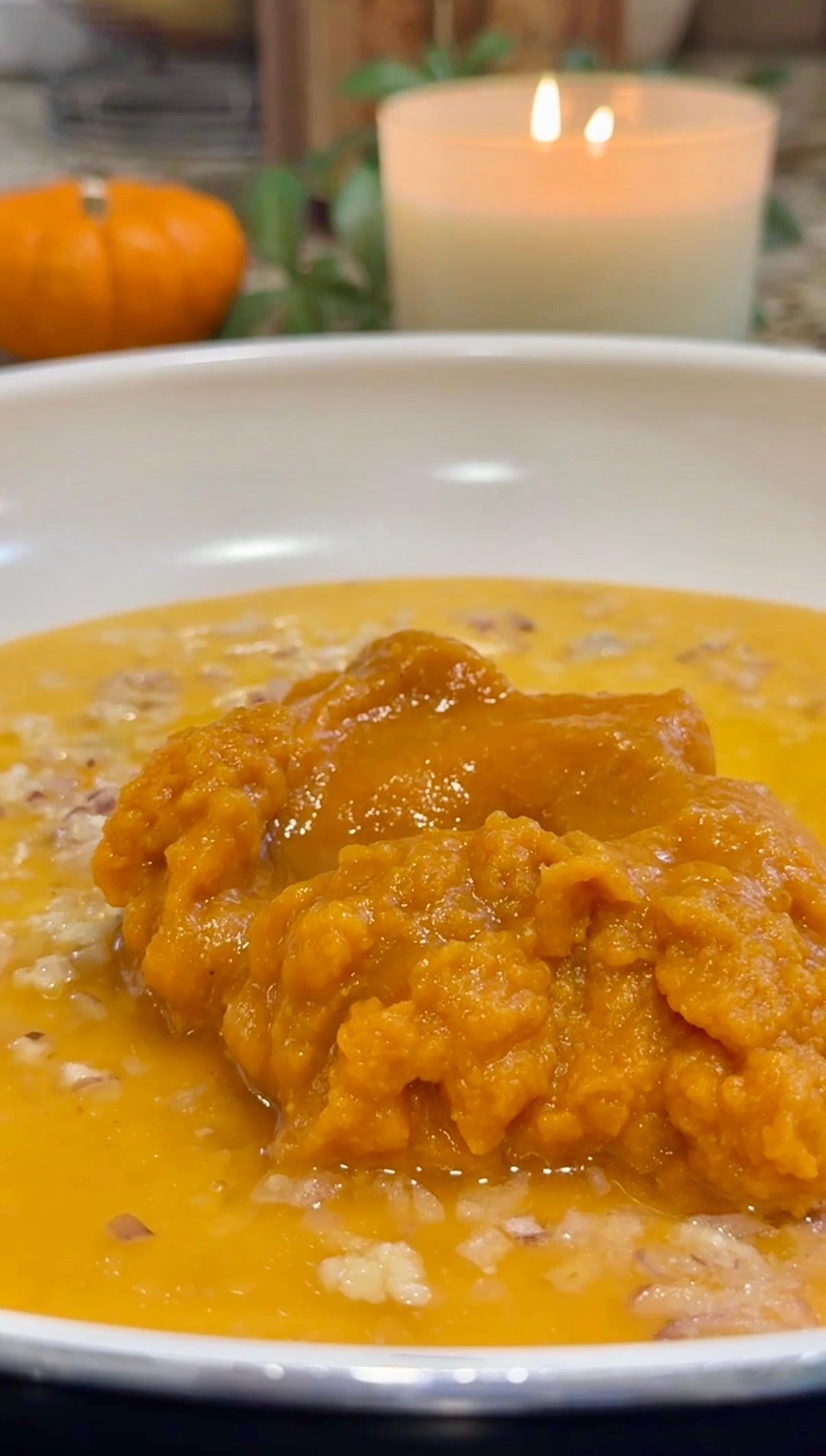 A close-up of a bowl with oatmeal topped with a generous scoop of orange pumpkin puree, set on a table with blurred candles and a pumpkin in the background.