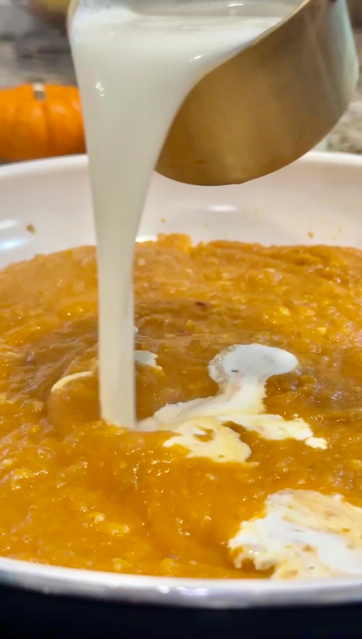 A close-up of cream being poured from a metal cup into a pan of orange pumpkin soup, with a small pumpkin visible in the background.