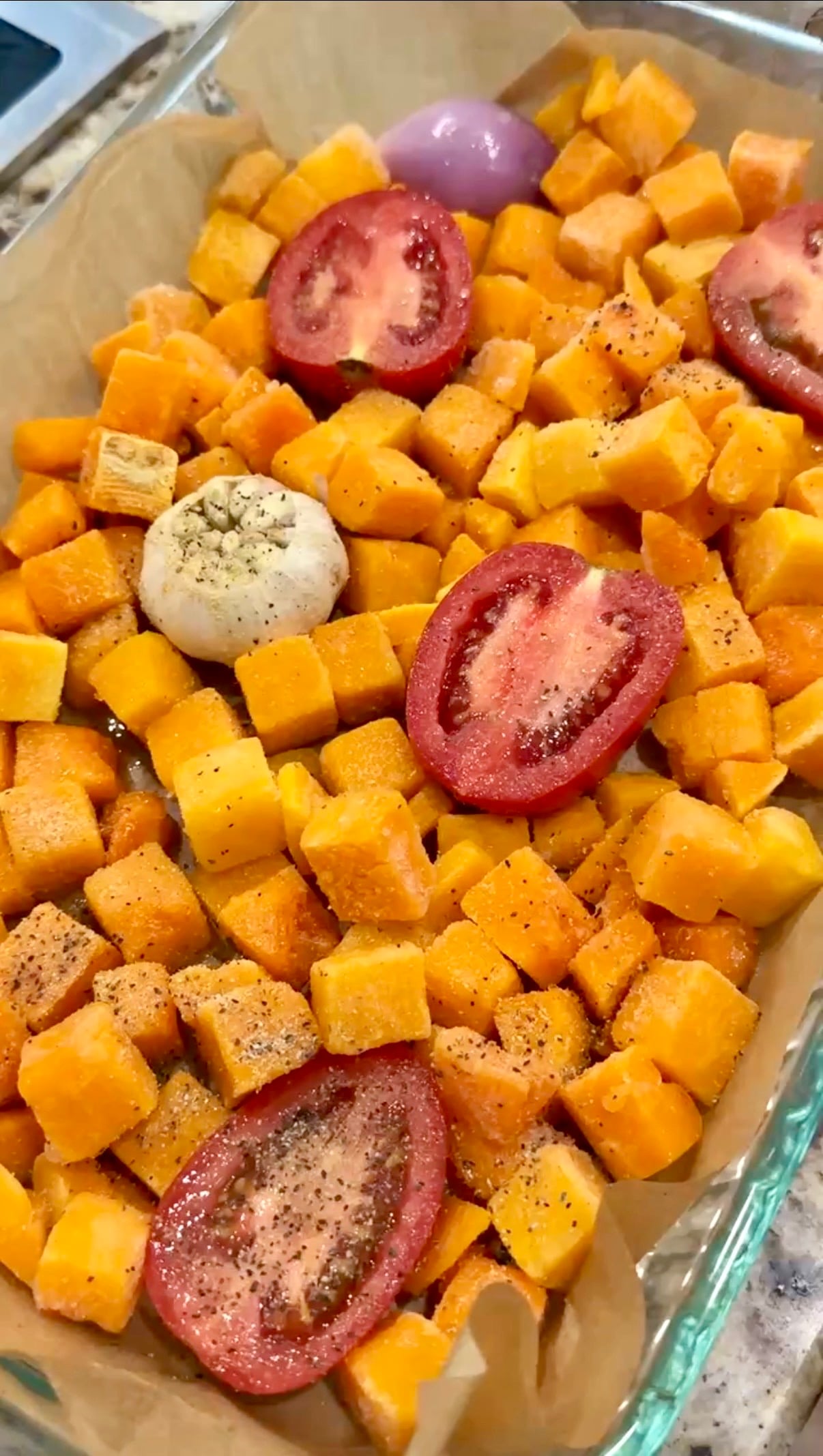 Diced butternut squash, halved tomatoes, a whole garlic bulb, and a halved red onion are seasoned and arranged in a glass baking dish, ready to be roasted.