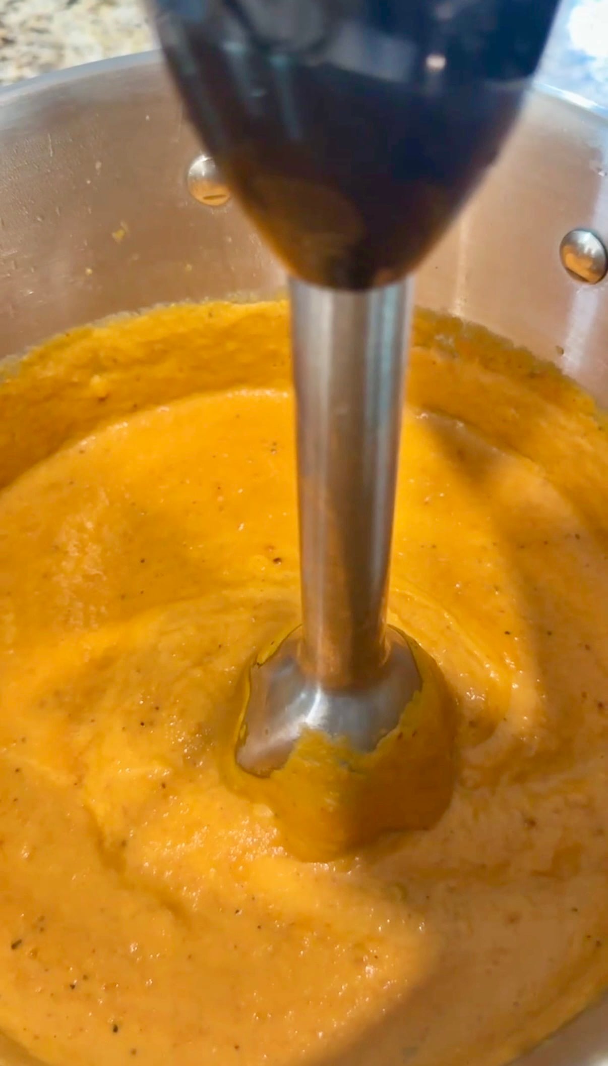 An immersion blender is blending thick orange soup in a metal pot, creating a smooth texture.