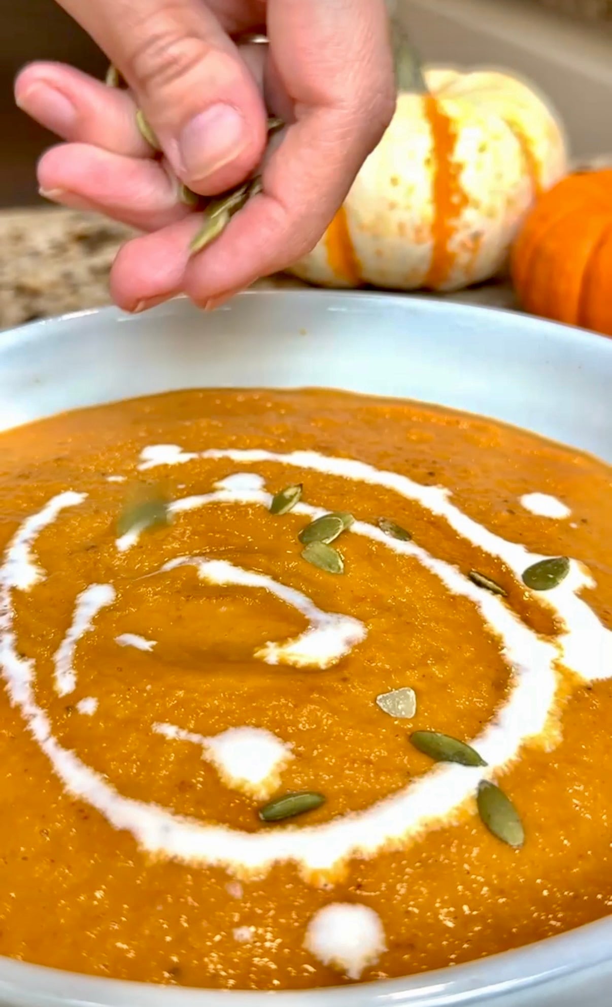A hand sprinkles pumpkin seeds on creamy orange soup with a swirl of cream. Two pumpkins are blurred in the background.