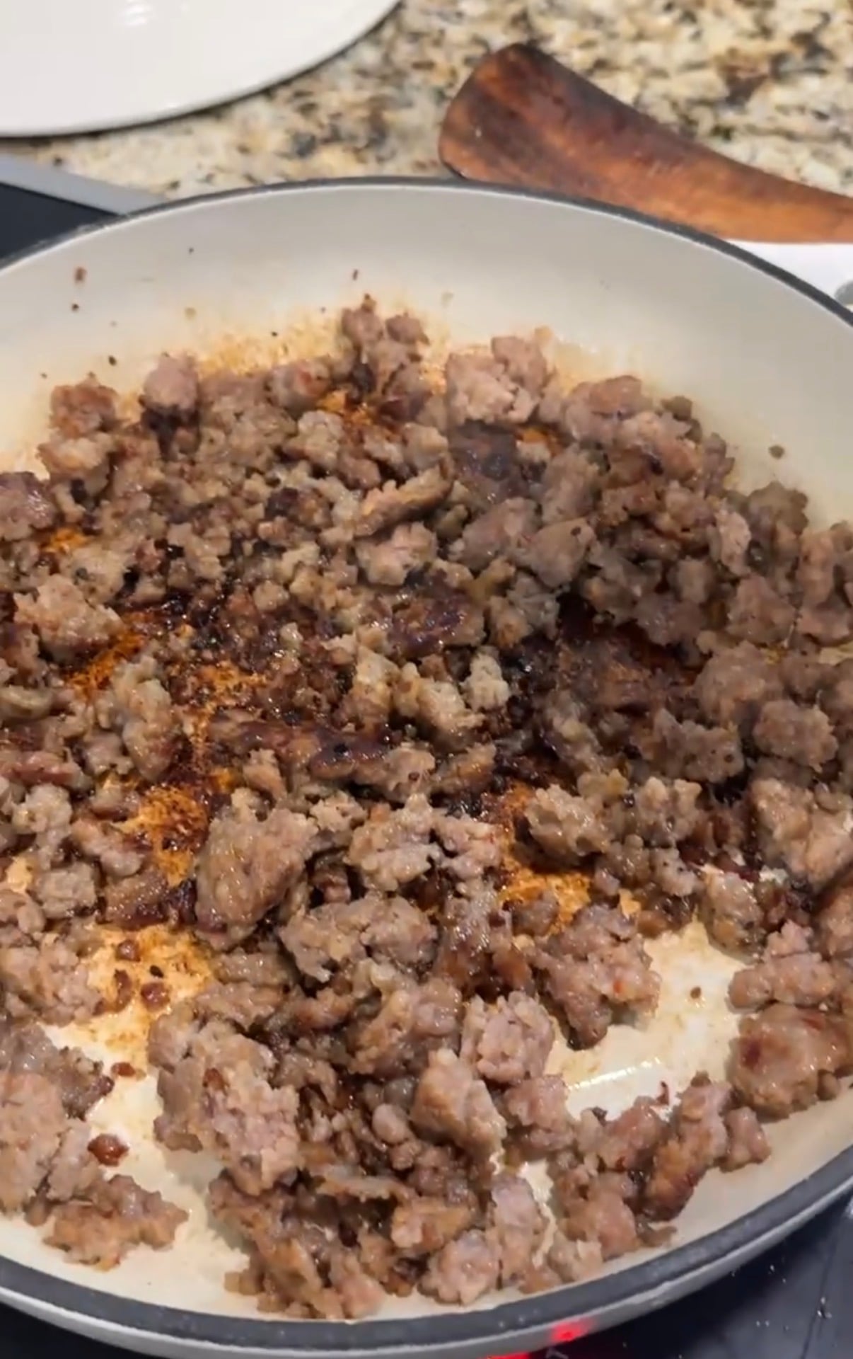 Ground sausage browning in a white skillet on a stovetop, with some areas caramelized. A wooden spatula and granite countertop are visible in the background.