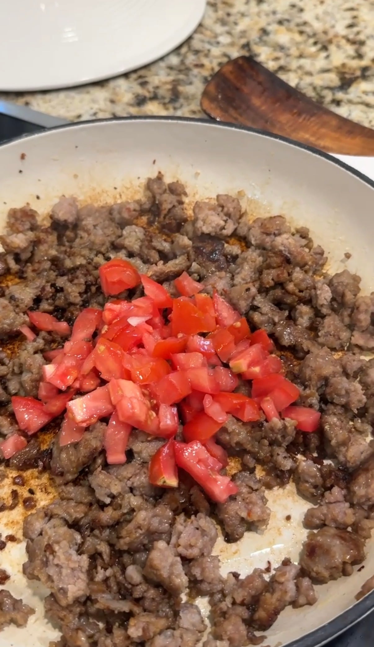 Ground meat is browning in a skillet with a pile of freshly chopped tomatoes added on top, ready to be mixed in and cooked together.