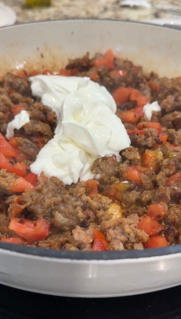 Ground meat is browning in a skillet with a pile of freshly chopped tomatoes added on top, ready to be mixed in and cooked together.