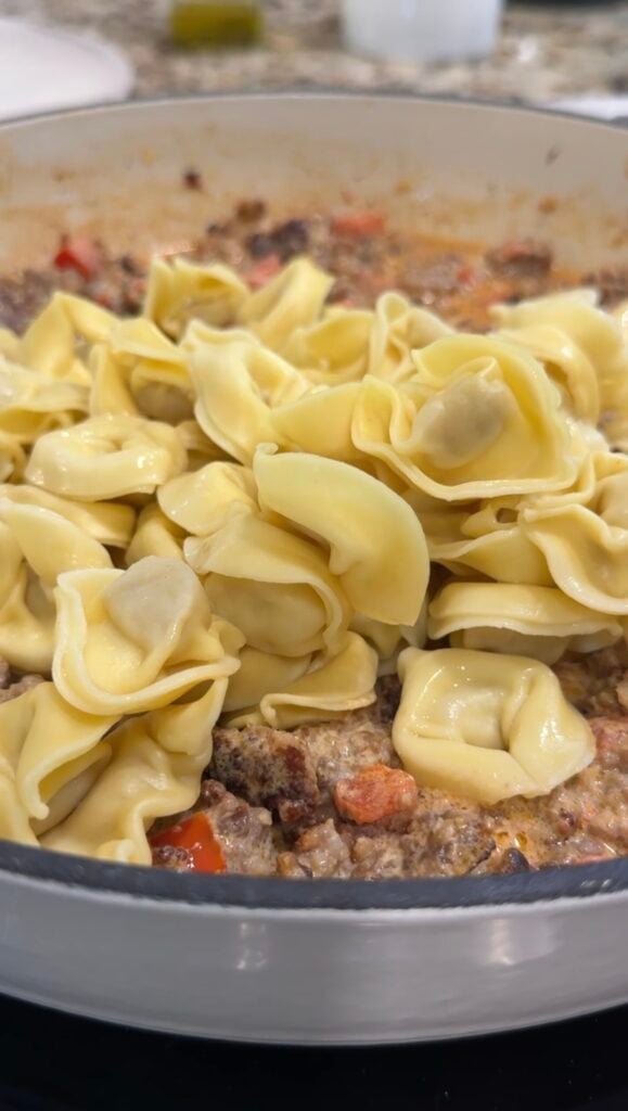 A close-up of uncooked tortellini pasta placed on top of a creamy mixture with ground meat and diced vegetables in a pan.