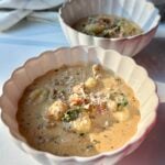 Two white scalloped bowls filled with creamy soup containing gnocchi, greens, and ground meat, garnished with grated cheese, sit on a white surface in warm natural light.