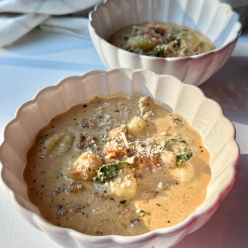 Two white scalloped bowls filled with creamy soup containing gnocchi, greens, and ground meat, garnished with grated cheese, sit on a white surface in warm natural light.