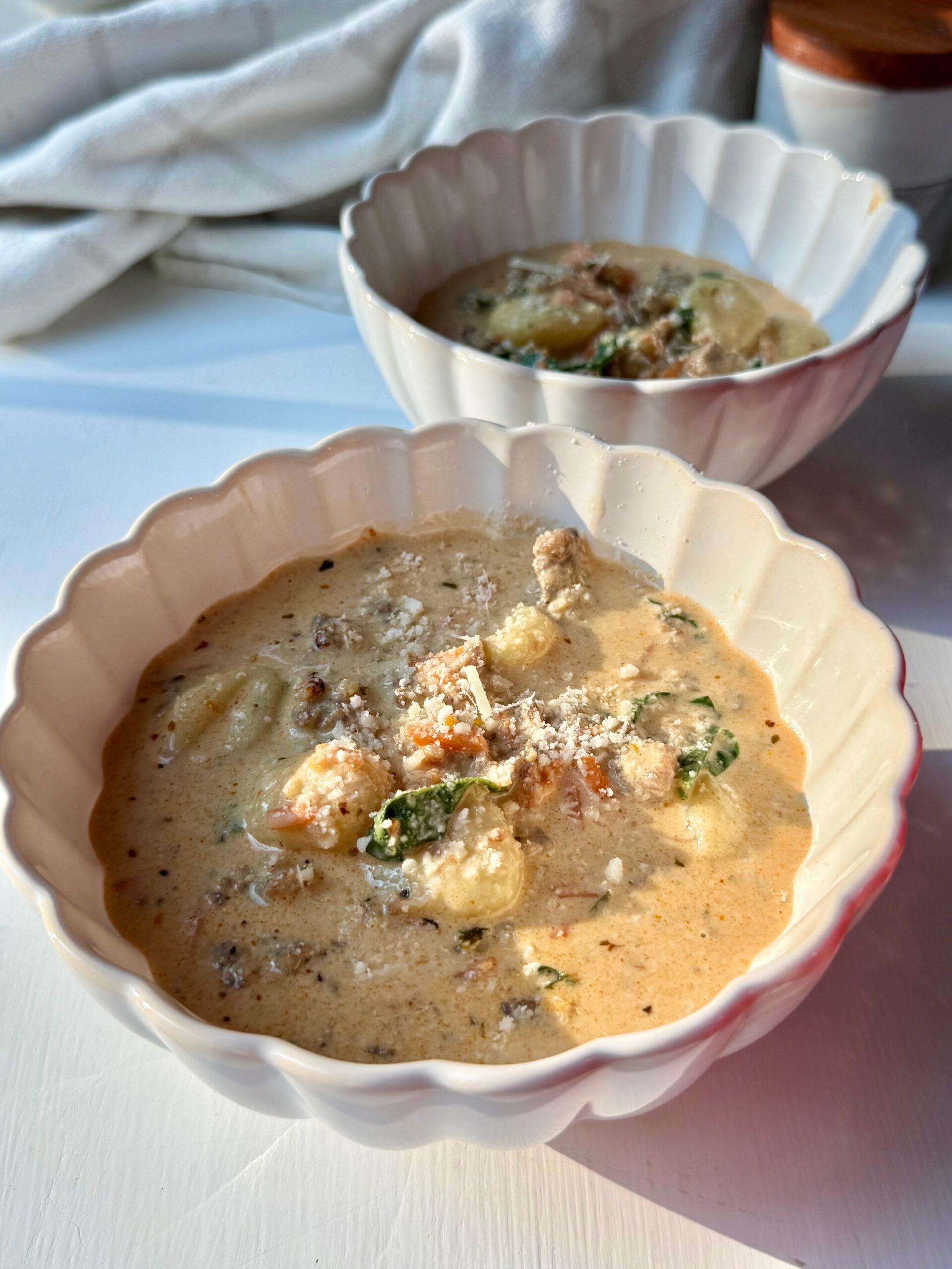 Two white scalloped bowls filled with creamy soup containing gnocchi, greens, and ground meat, garnished with grated cheese, sit on a white surface in warm natural light.