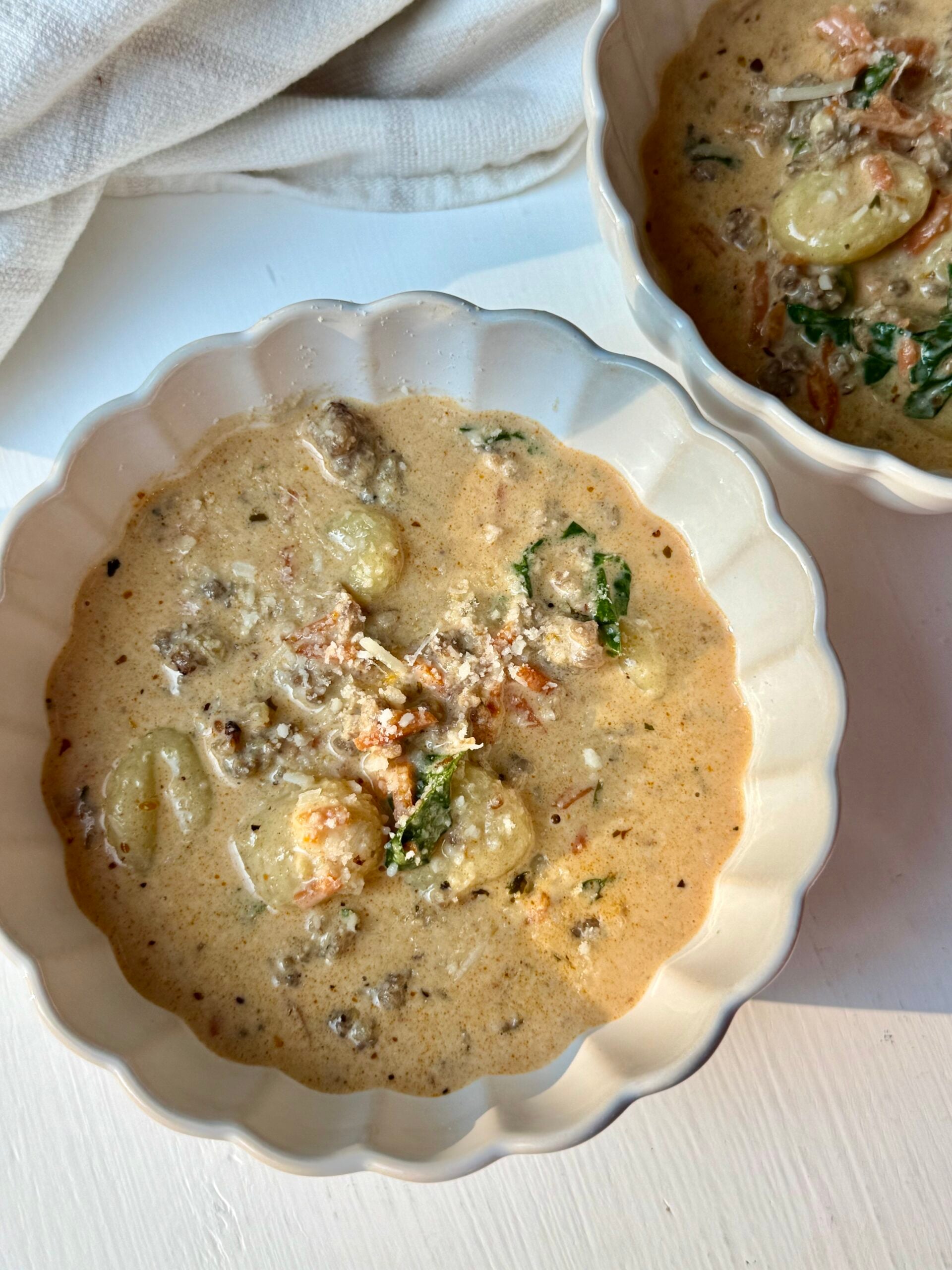Two bowls of creamy soup with gnocchi, spinach, ground meat, and grated cheese are placed on a white surface next to a beige cloth.