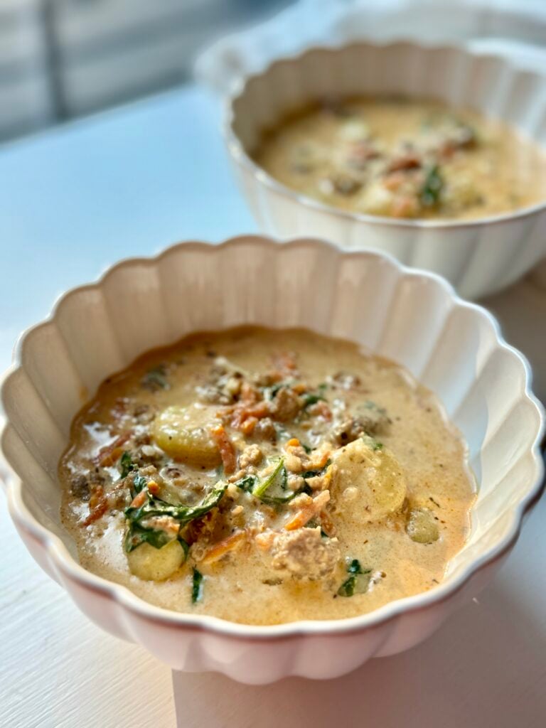 Two white scalloped bowls filled with creamy soup containing gnocchi, greens, and ground meat, garnished with grated cheese, sit on a white surface in warm natural light.