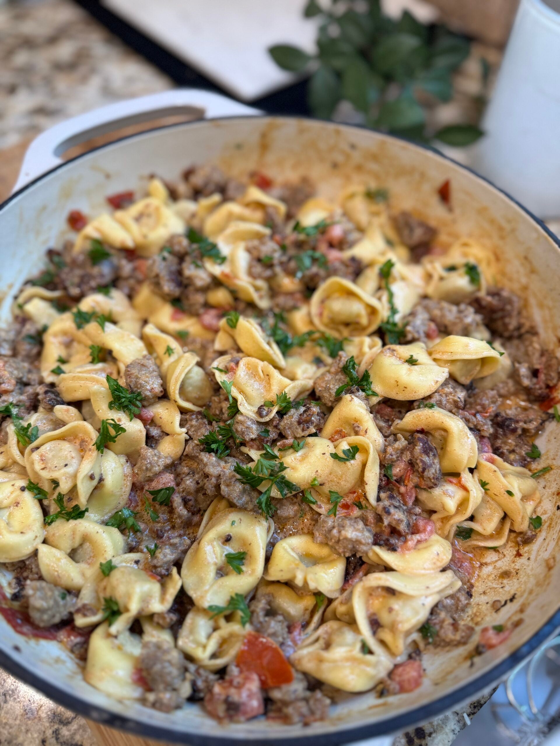 A skillet filled with cheesy tortellini pasta, ground sausage, chopped tomatoes, and topped with fresh parsley, all mixed in a creamy sauce.