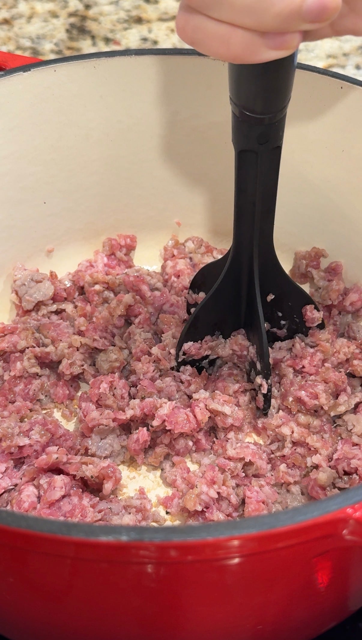 Ground meat is being broken up and cooked in a white pot with a black meat chopper. Some pieces are browned, while others are still pink. The edge of the red pot is visible at the bottom of the image.