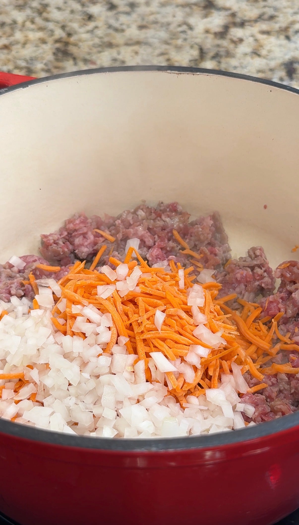 Chopped onions, shredded carrots, and ground meat are being cooked together in a large white pot on a stovetop.