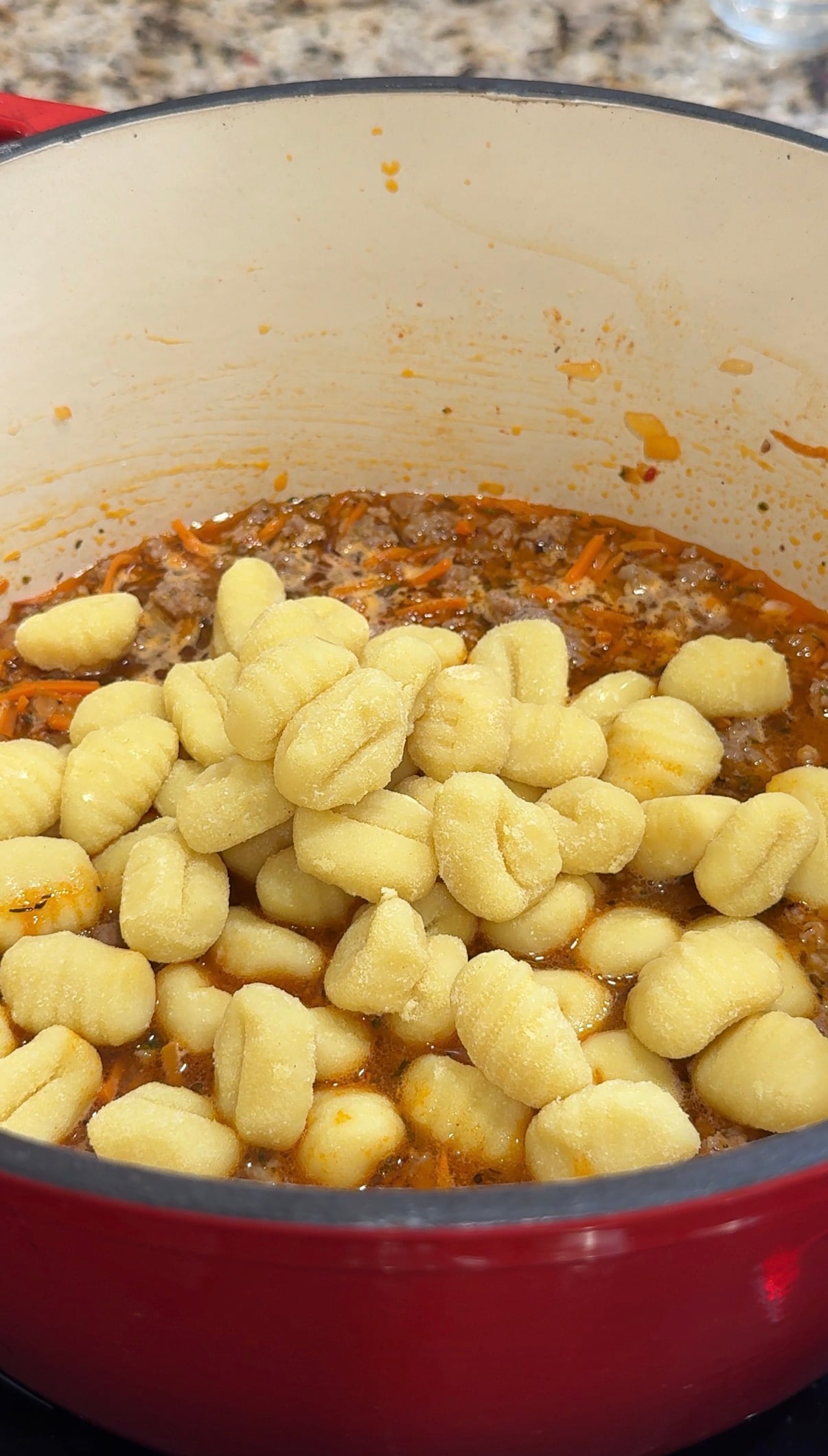 Fresh gnocchi is piled on top of a simmering meat and vegetable sauce in a red pot, ready to be mixed and cooked together.