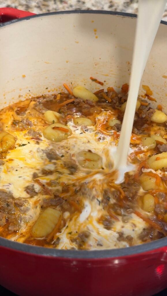 Cream is being poured into a pot of soup with ground meat, gnocchi, and vegetables, creating a marbled effect as it mixes with the broth.