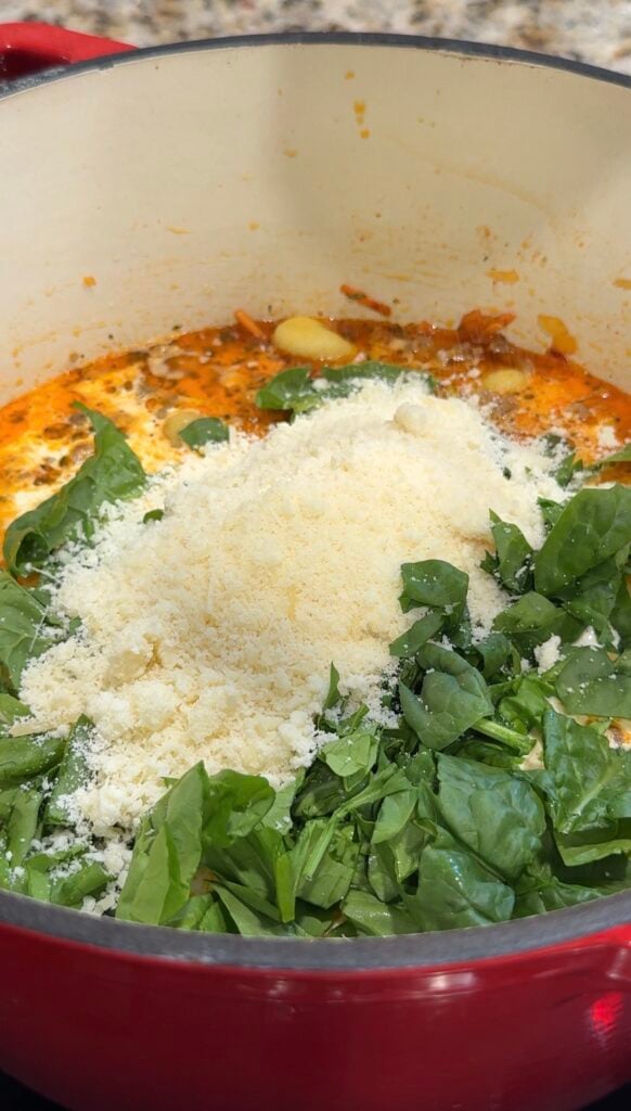 Chopped spinach and grated cheese are added to a pot of soup, with visible broth and onions. The mixture appears to be cooking, with vibrant green and orange colors.
