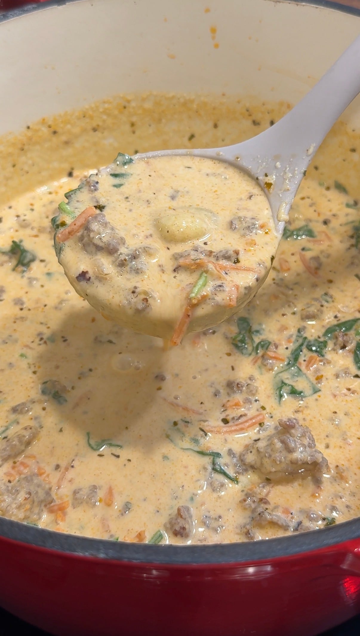 A close-up of a creamy soup with sausage, spinach, shredded carrots, and seasonings. A ladle lifts a portion from the pot, showing chunky and colorful ingredients in the rich broth.