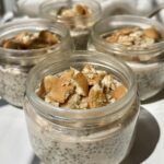 Four small glass jars filled with chia pudding and topped with crushed vanilla wafer cookies are arranged on a white surface in bright natural light.