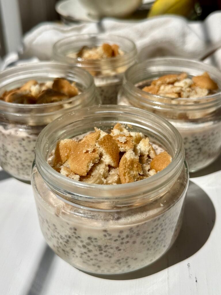 Four small glass jars filled with chia pudding and topped with crushed vanilla wafer cookies are arranged on a white surface in bright natural light.