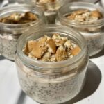 Four small glass jars filled with chia pudding and topped with crushed vanilla wafer cookies are arranged on a white surface in bright natural light.