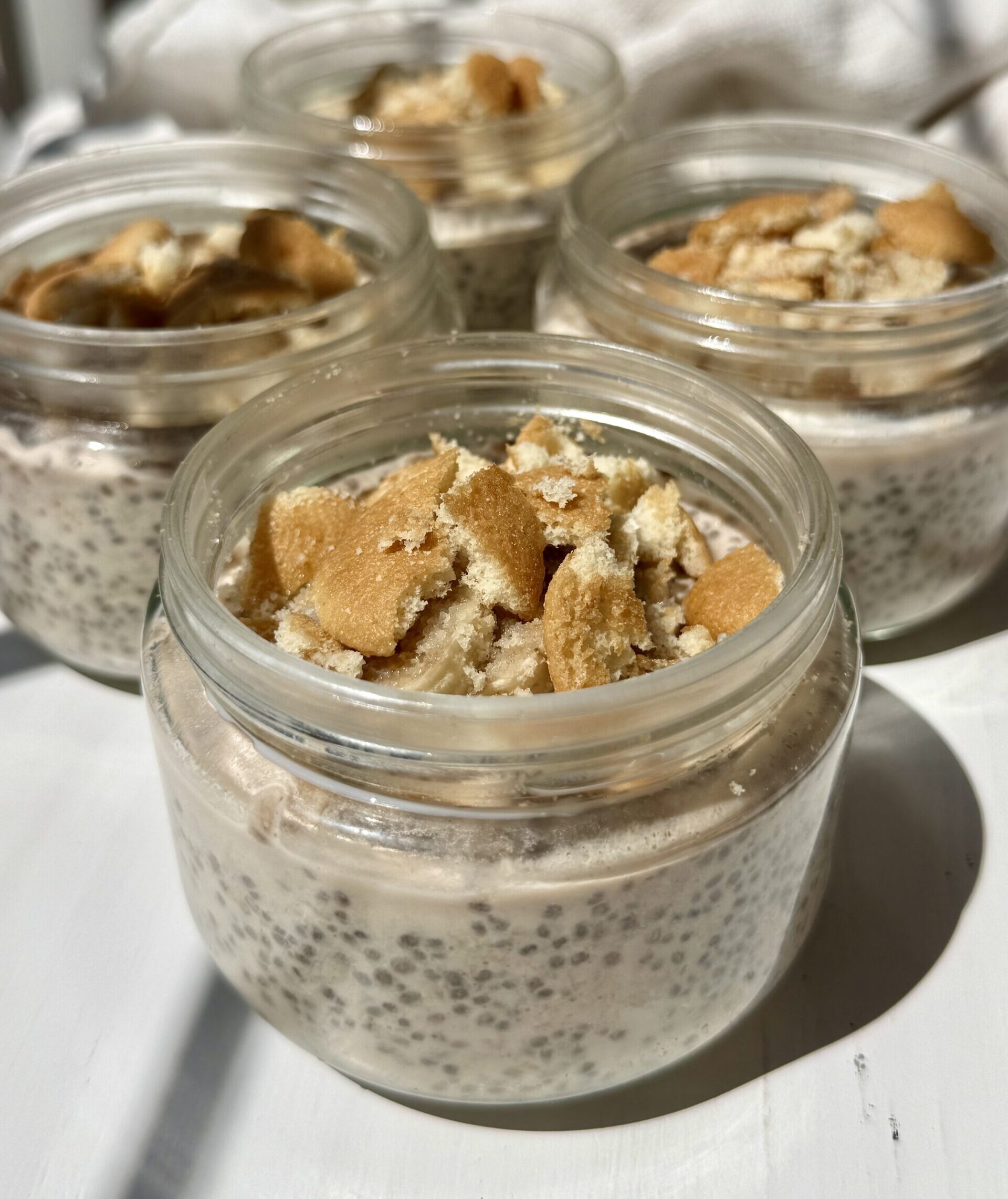 Four small glass jars filled with chia pudding and topped with crushed vanilla wafer cookies are arranged on a white surface in bright natural light.