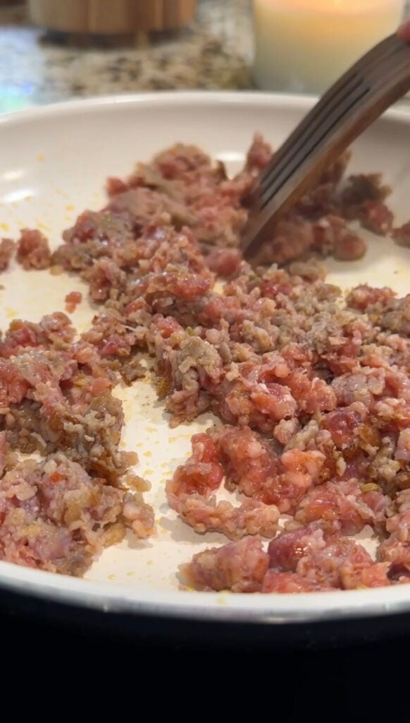 Ground beef is being cooked in a white skillet, with some meat still pink and other portions browned. A utensil is visible stirring or breaking up the meat.