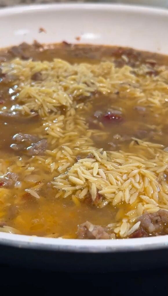 Close-up of orzo pasta simmering in a broth with bits of ground meat and small pieces of tomato visible in a pan.