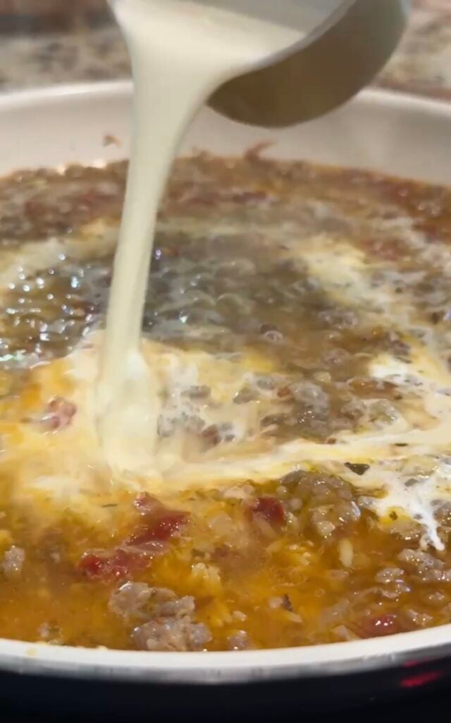 Cream is being poured into a bubbling mixture of ground meat, tomatoes, and oil in a pan, likely as part of cooking a sauce.