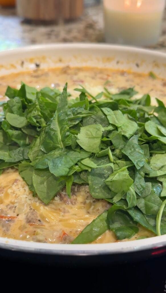 A creamy pasta dish in a pan is topped with fresh spinach leaves, ready to be stirred in.