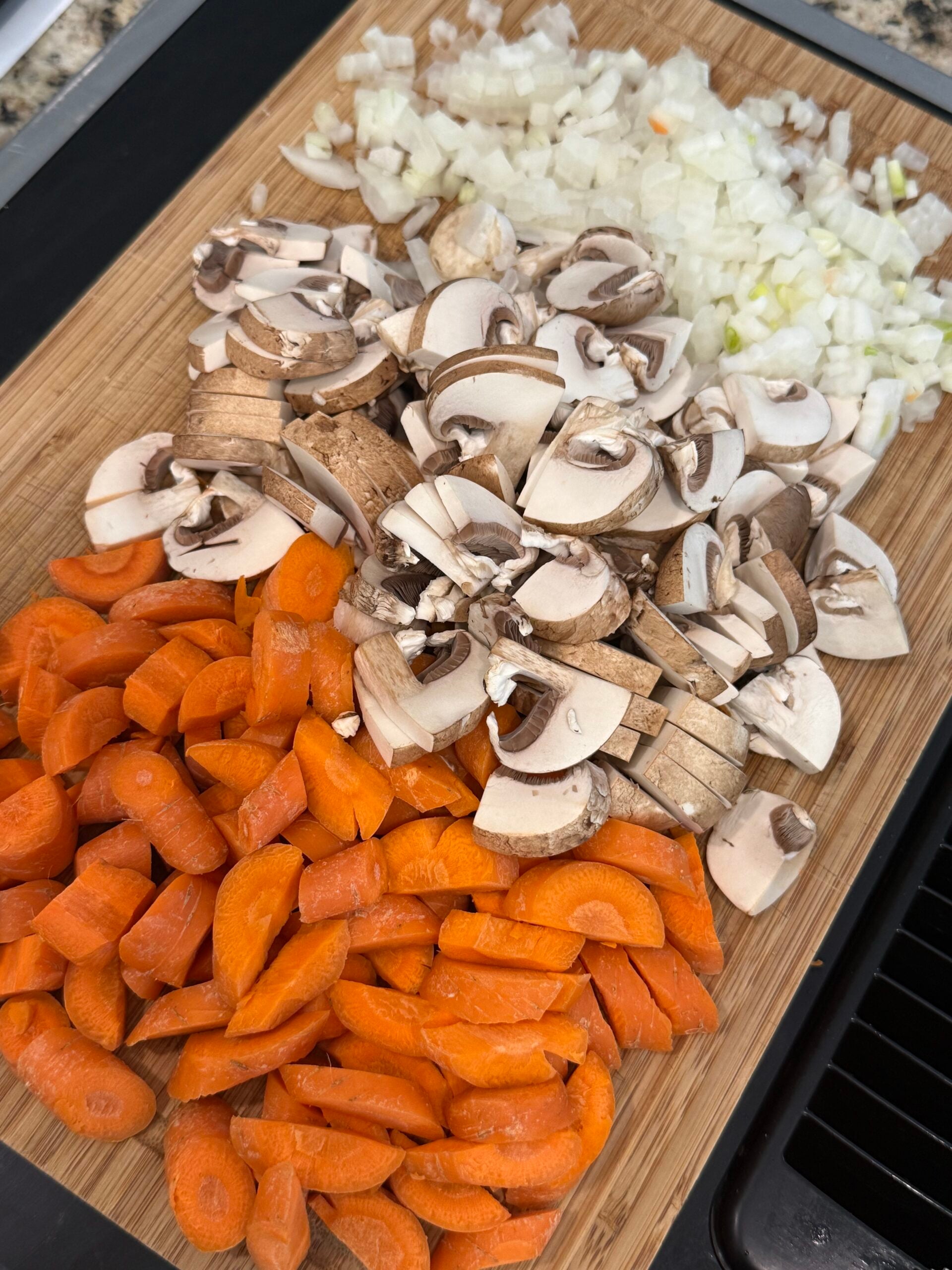 Chopped carrots, mushrooms, and onions arranged neatly on a wooden cutting board, ready for cooking.