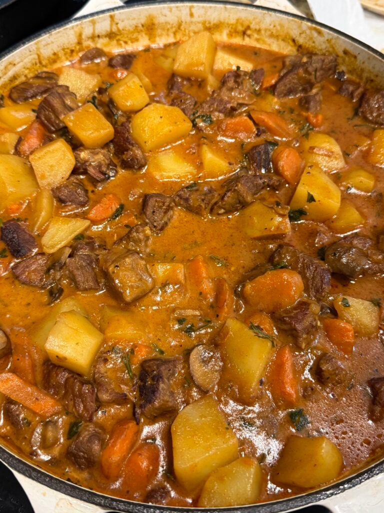 A close-up of a hearty beef stew in a pot, filled with chunks of beef, potatoes, carrots, and mushrooms in a rich brown broth.