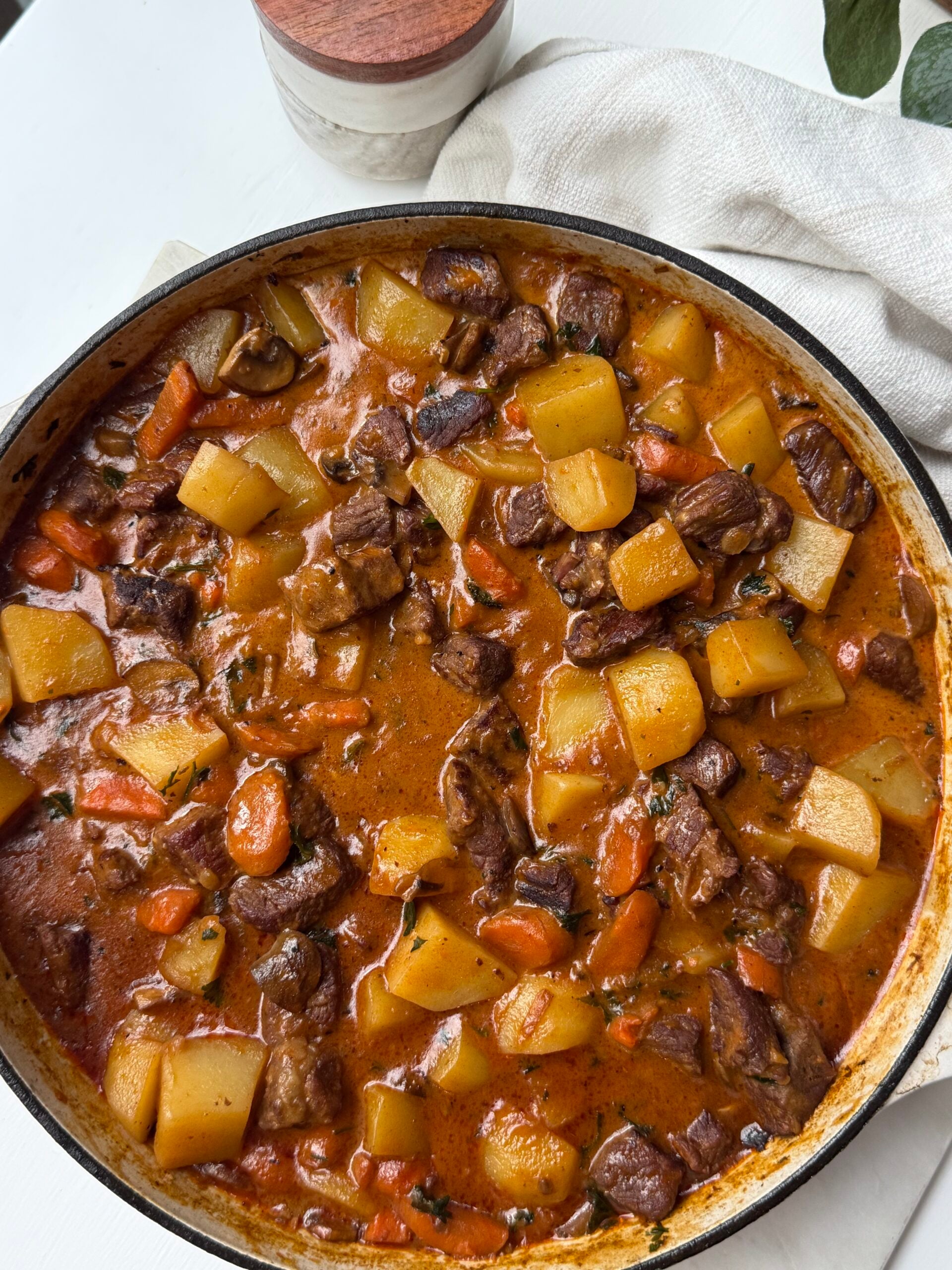 A pot of hearty beef stew with chunks of beef, potatoes, carrots, and herbs in a rich brown broth, sitting on a white surface beside a napkin and a small container.
