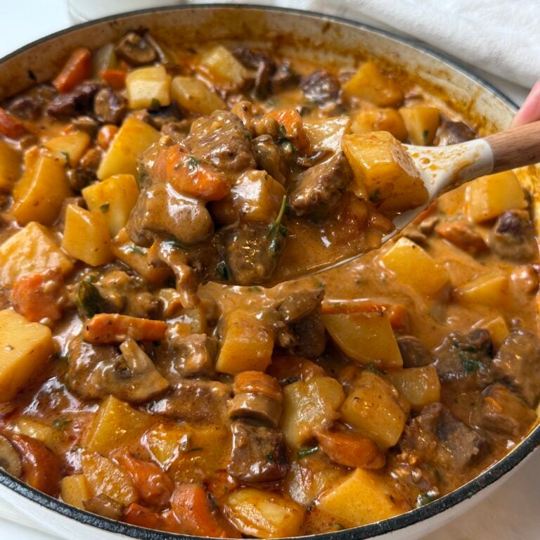A close-up of a hearty beef stew with chunks of beef, potatoes, carrots, and herbs in a thick, rich sauce. A wooden spoon is lifting a serving from the pot.