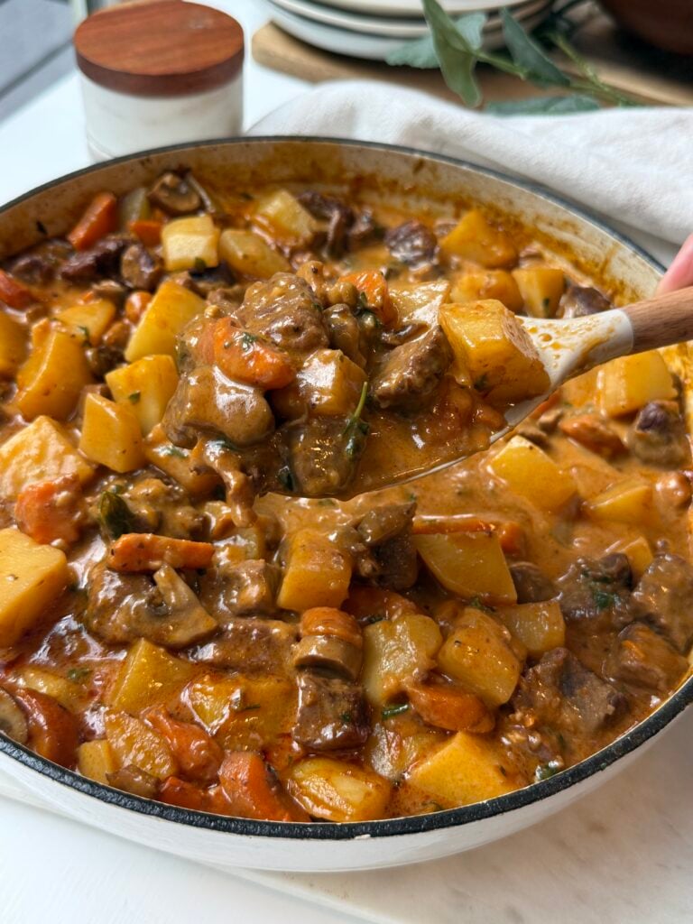 A wooden spoon scoops hearty beef stew with chunks of beef, carrots, potatoes, and rutabaga in a creamy broth from a large pot on a white surface.