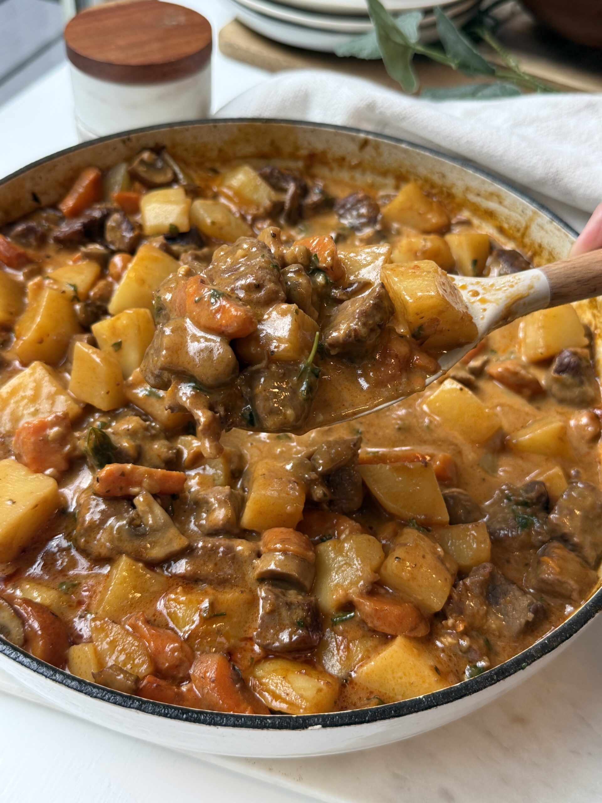 A wooden spoon scoops hearty beef stew with chunks of beef, carrots, potatoes, and rutabaga in a creamy broth from a large pot on a white surface.
