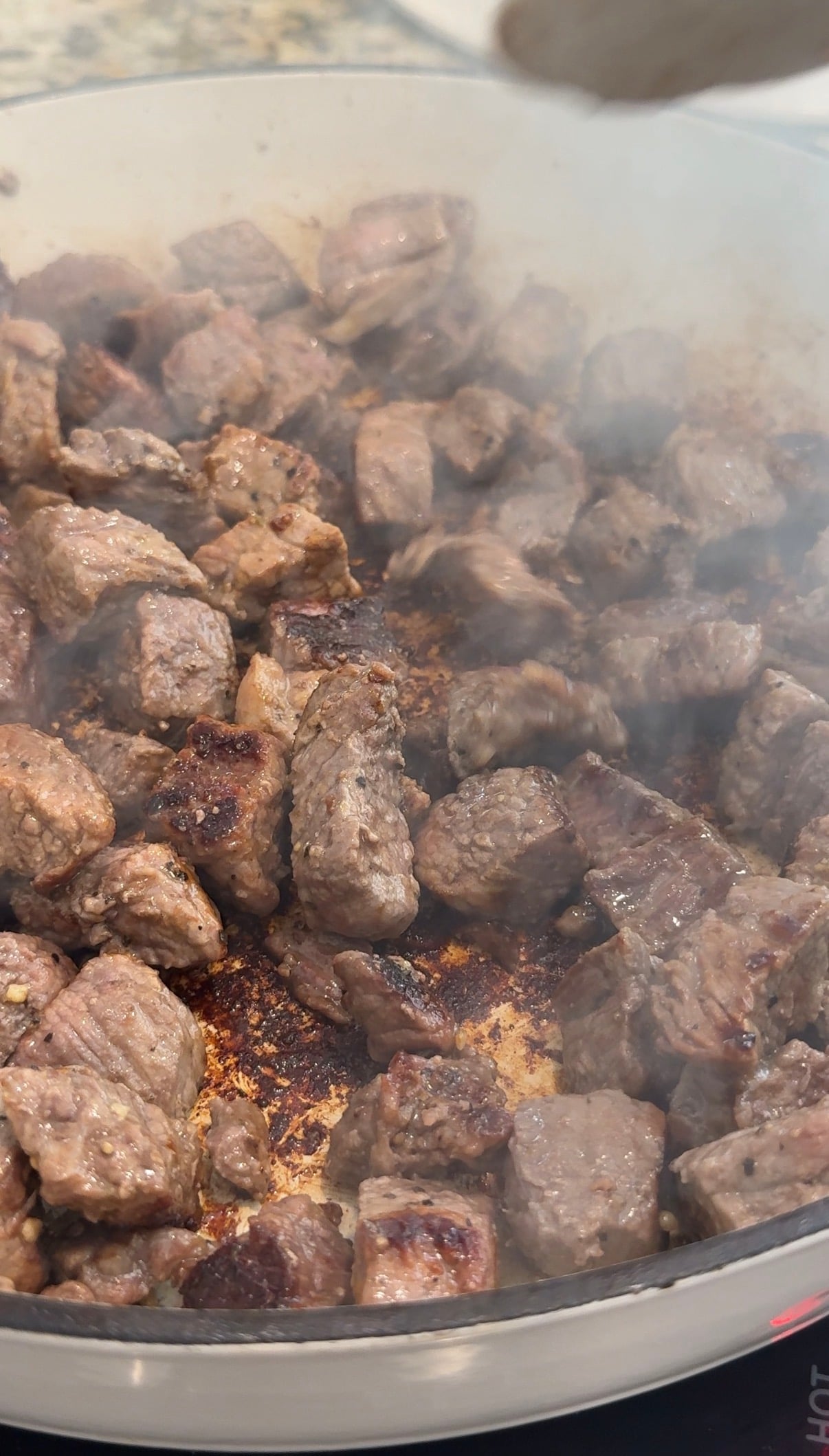 Chunks of beef are browning in a pan, with some pieces showing a seared crust. Steam or smoke rises, indicating the meat is being cooked at high heat.