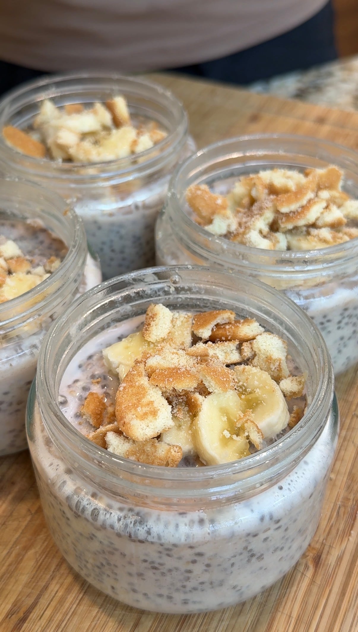 Four glass jars filled with chia pudding topped with banana slices and crumbled cookies are arranged on a wooden surface.