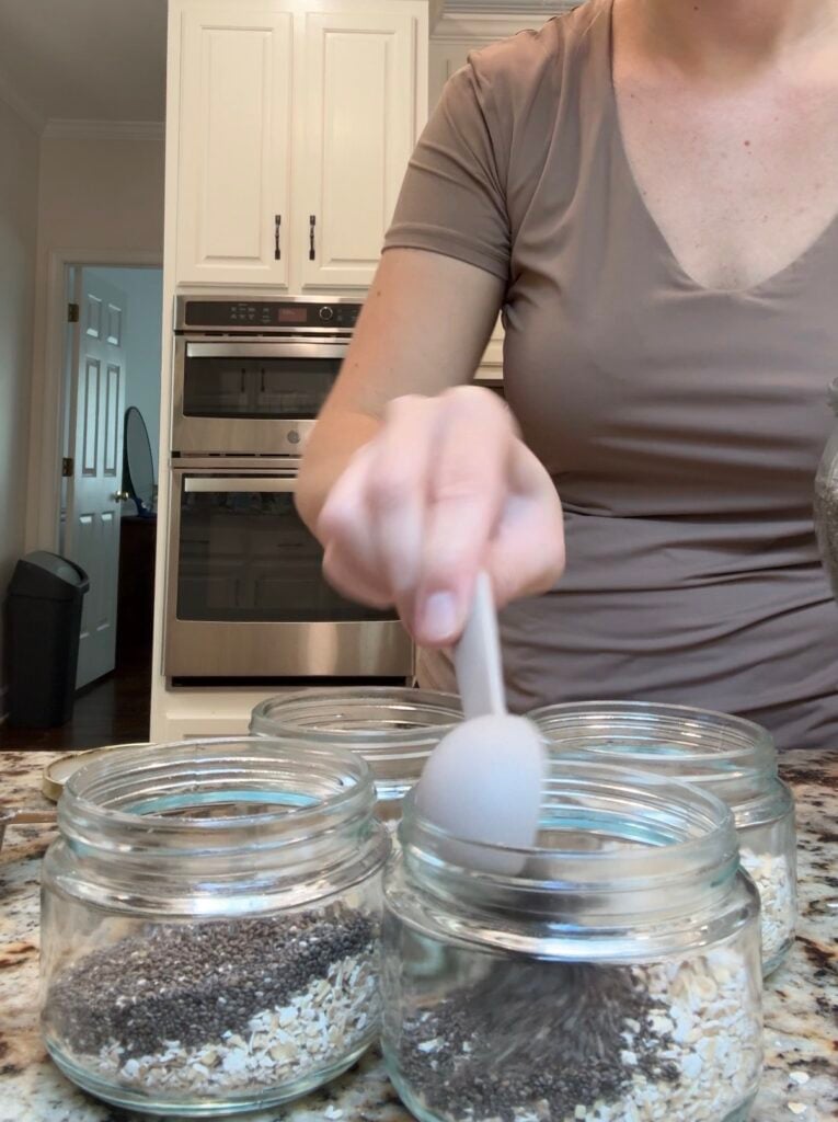 A person fills glass jars with oats and chia seeds using a spoon on a kitchen counter; kitchen cabinets and an oven are visible in the background.