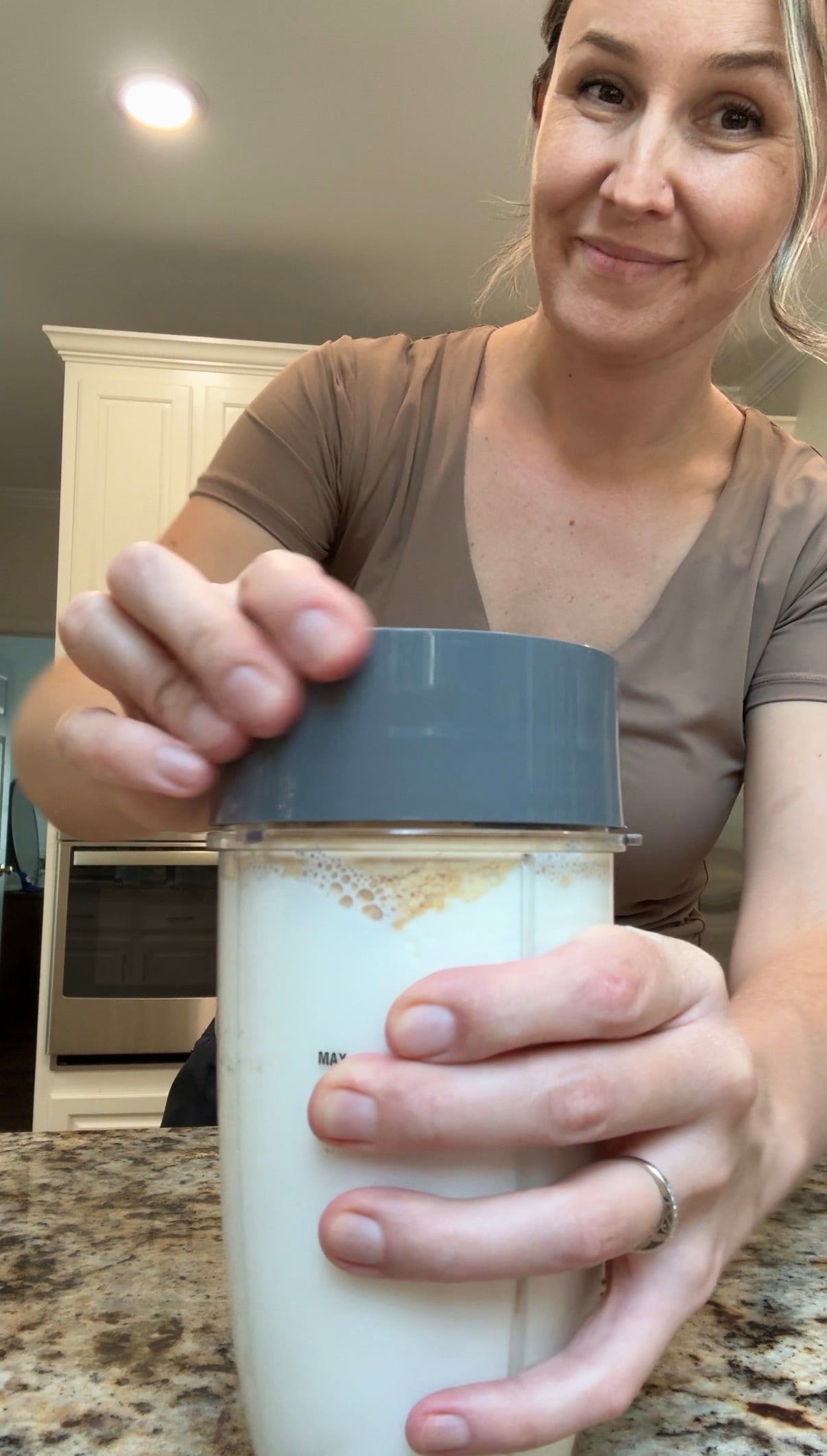 A woman smiles as she places a lid on a blender cup filled with milk, standing in a kitchen with white cabinets and granite countertop.