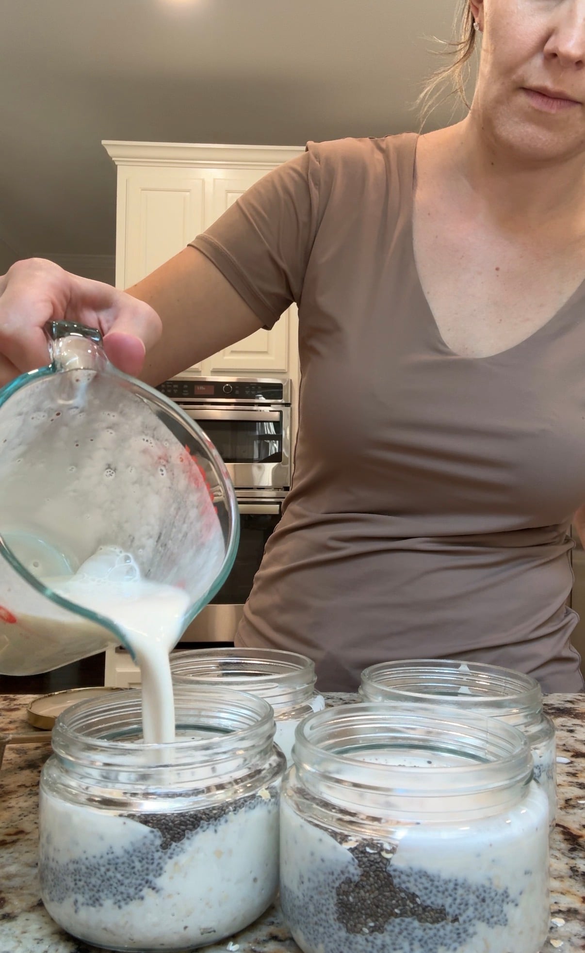 A woman pours milk from a measuring cup into glass jars containing chia seeds, preparing food on a kitchen counter.