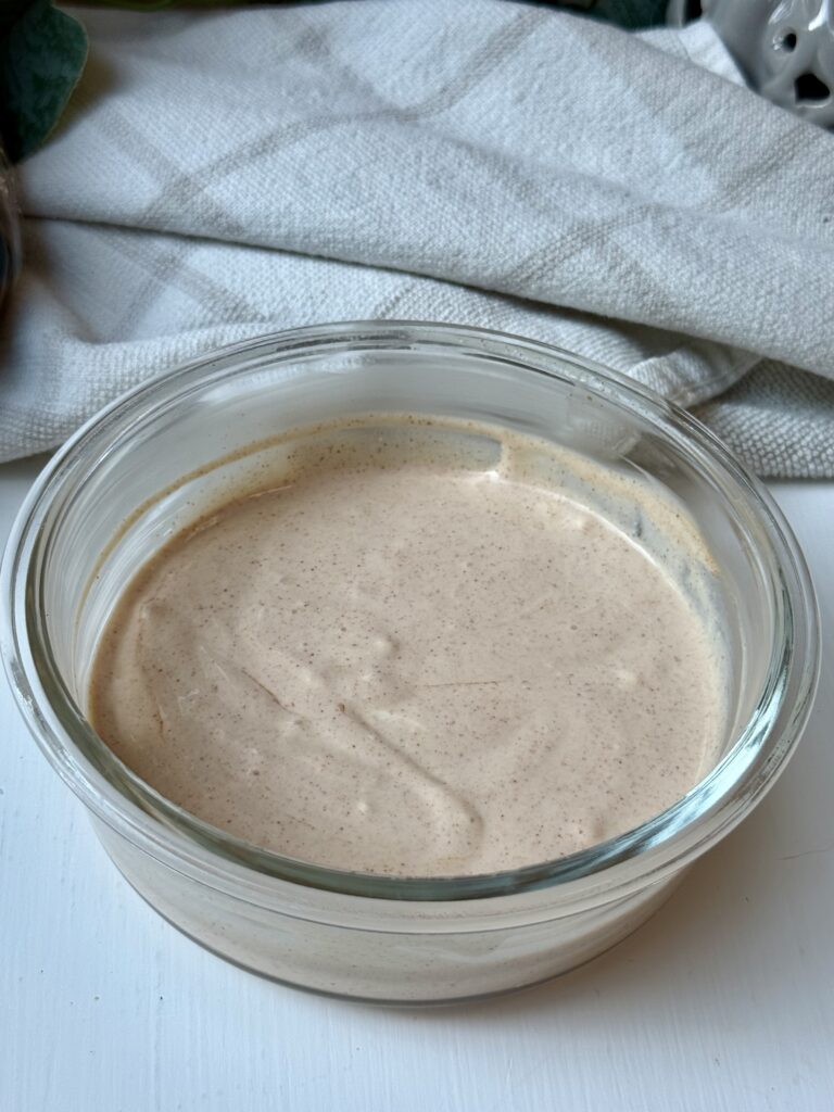 A glass bowl filled with creamy, light brown sauce sits on a white surface with a folded gray cloth in the background.