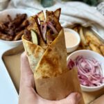 A hand holds a wrapped gyro filled with fries, grilled meat, pickles, onions, and creamy sauce. In the background are bowls of meat, red onions, sauce, and a tray of fries.